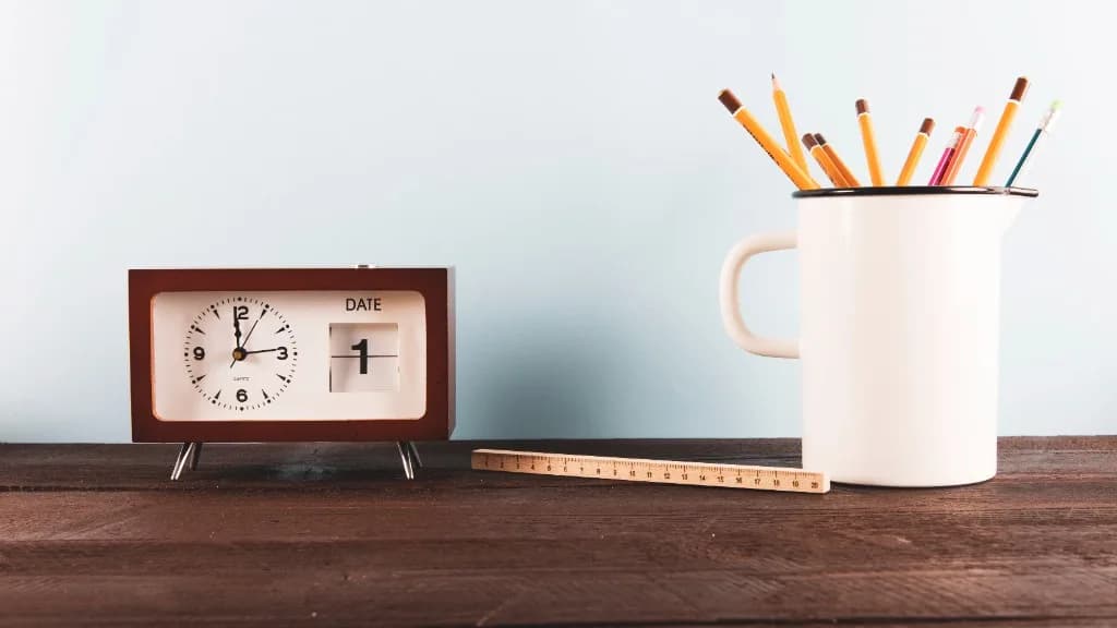 Reloj de escritorio, regla y lápices sobre una mesa de madera.
