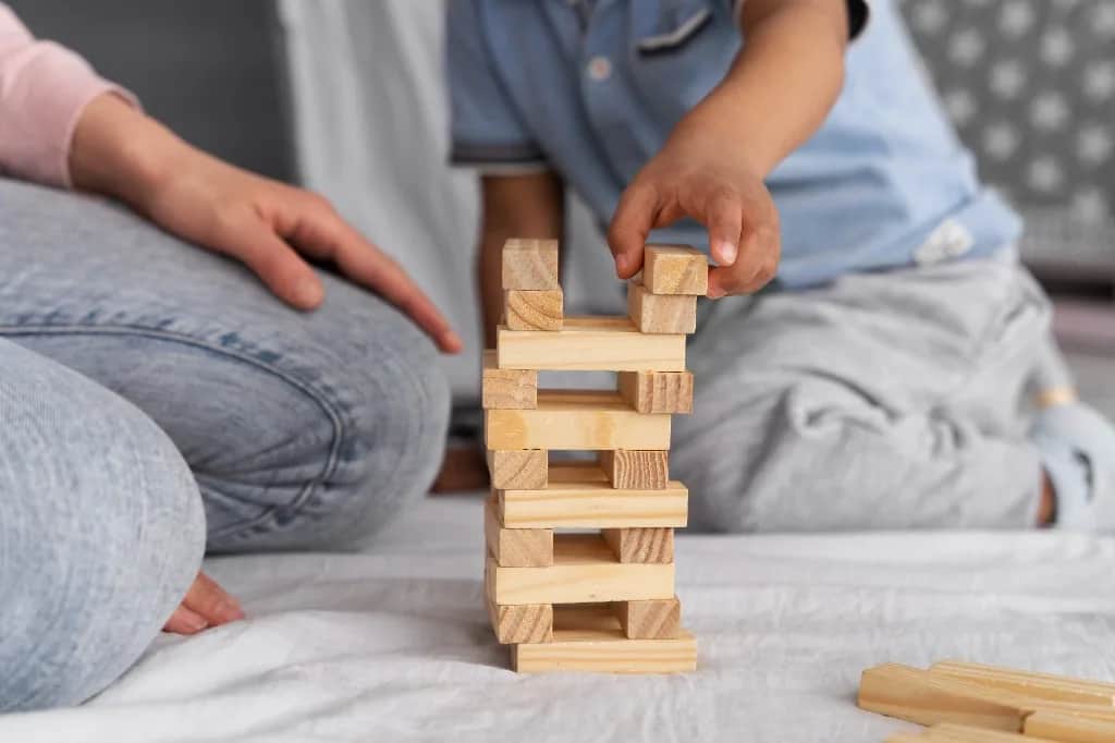 Mano de un niño colocando un bloque de madera en una torre, junto a un adulto.