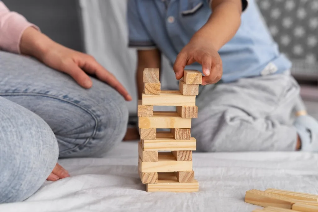 Mano de un niño colocando un bloque de madera en una torre, junto a un adulto.
