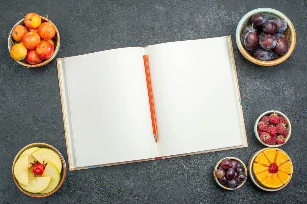 Cuaderno abierto y cuencos con fruta fresca vistos desde arriba.