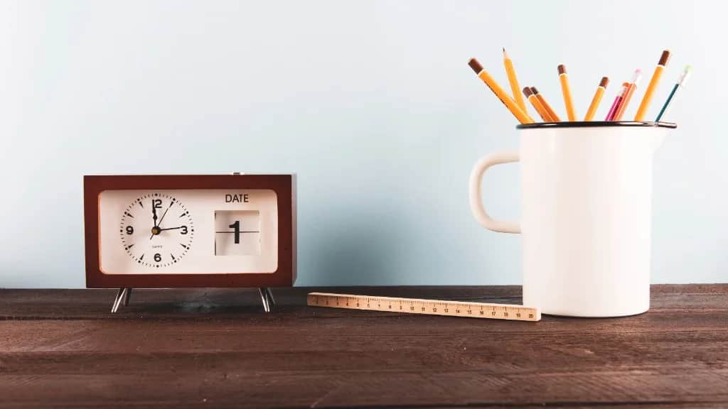 Reloj de escritorio, regla y lápices sobre una mesa de madera.